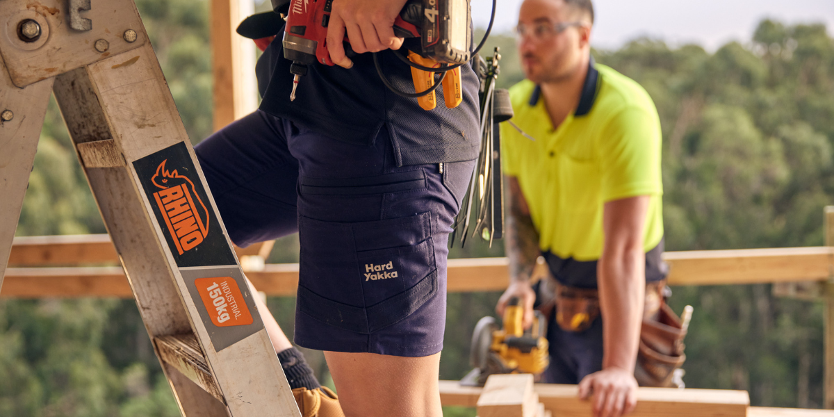 Person wearing Hard Yakka work shorts standing on a ladder, with another worker in the background.