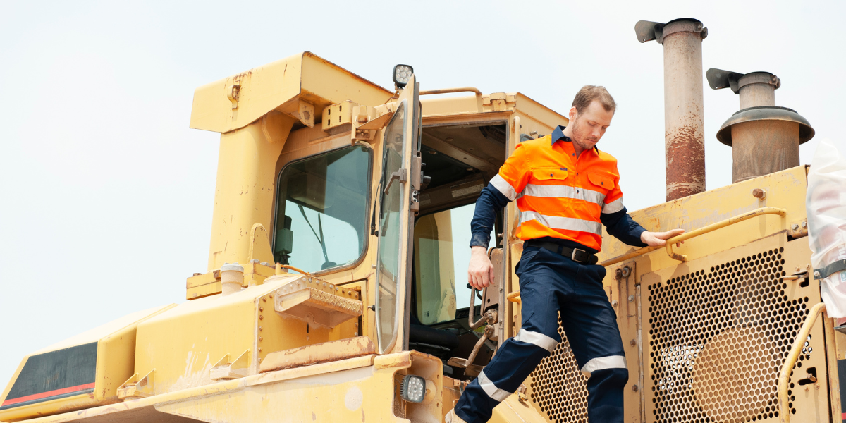 Person wearing an orange high-visibility jacket and pants stepping down from a large yellow construction vehicle.