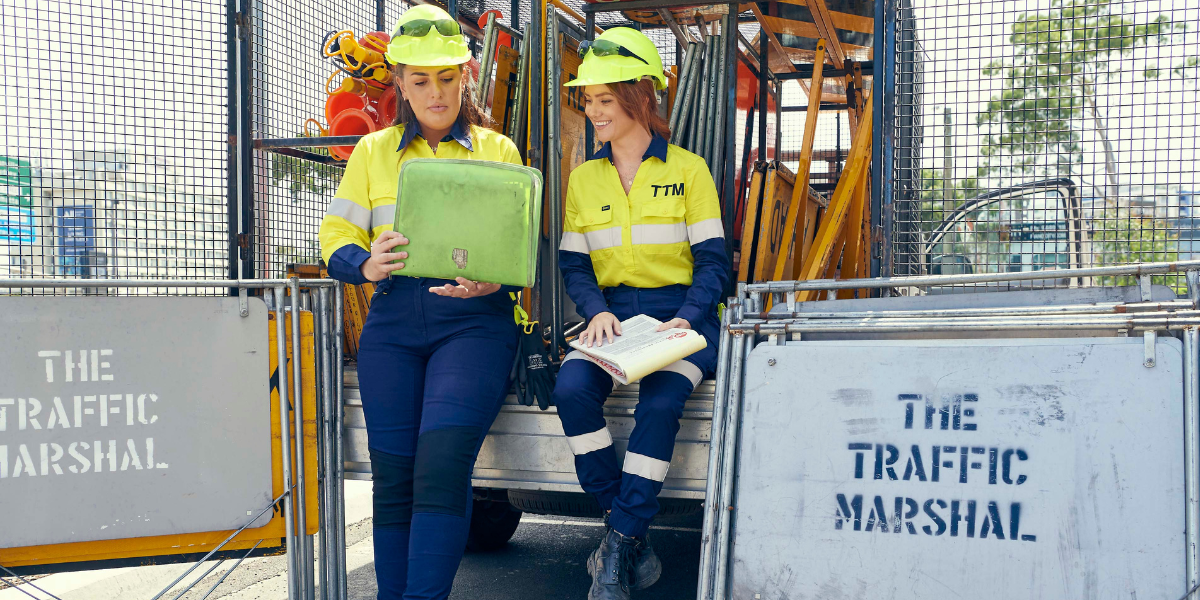 Two people wearing high-visibility uniforms and hard hats, sitting and reading on a construction site near a sign that says "The Traffic Marshal."