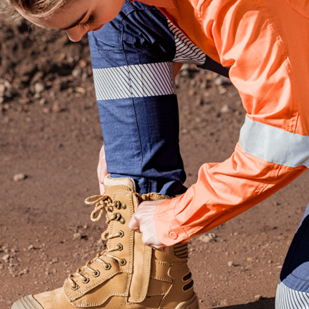 Person tying laces on tan work boots with blue pants and an orange high-visibility jacket.