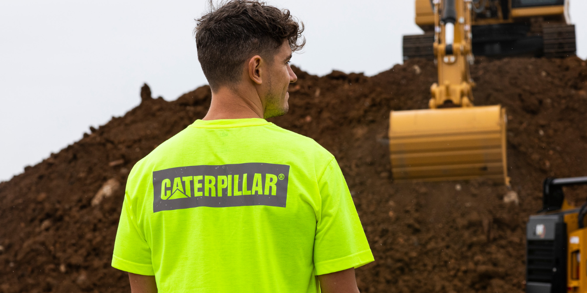 Person wearing a bright yellow Caterpillar t-shirt standing in front of construction equipment.